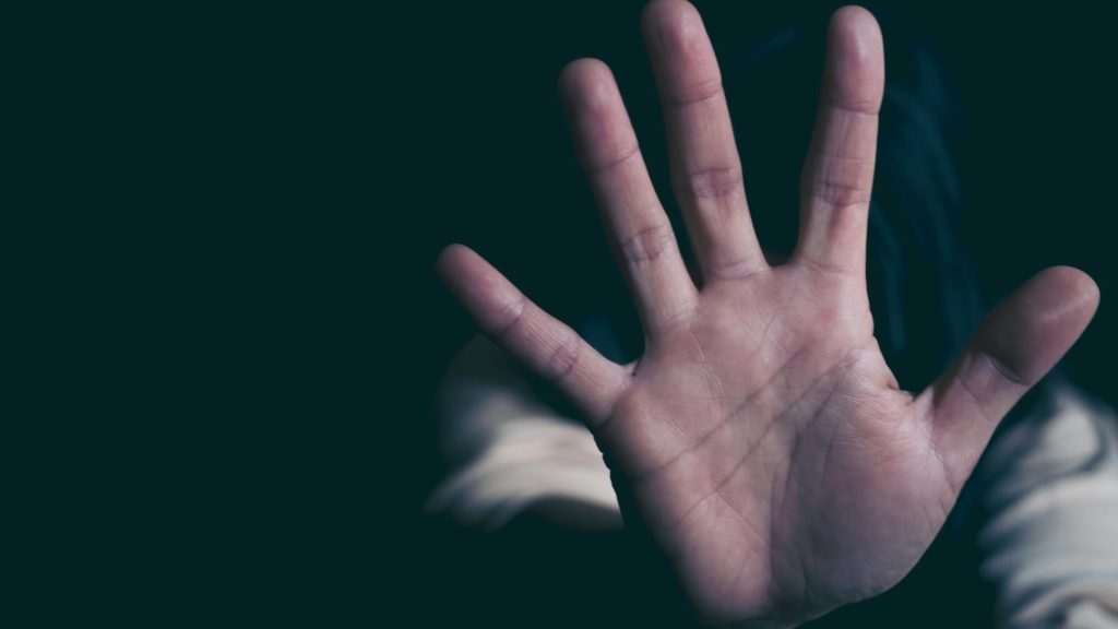 A woman huddled in the darkness with her hand in front of her signaling "stop"