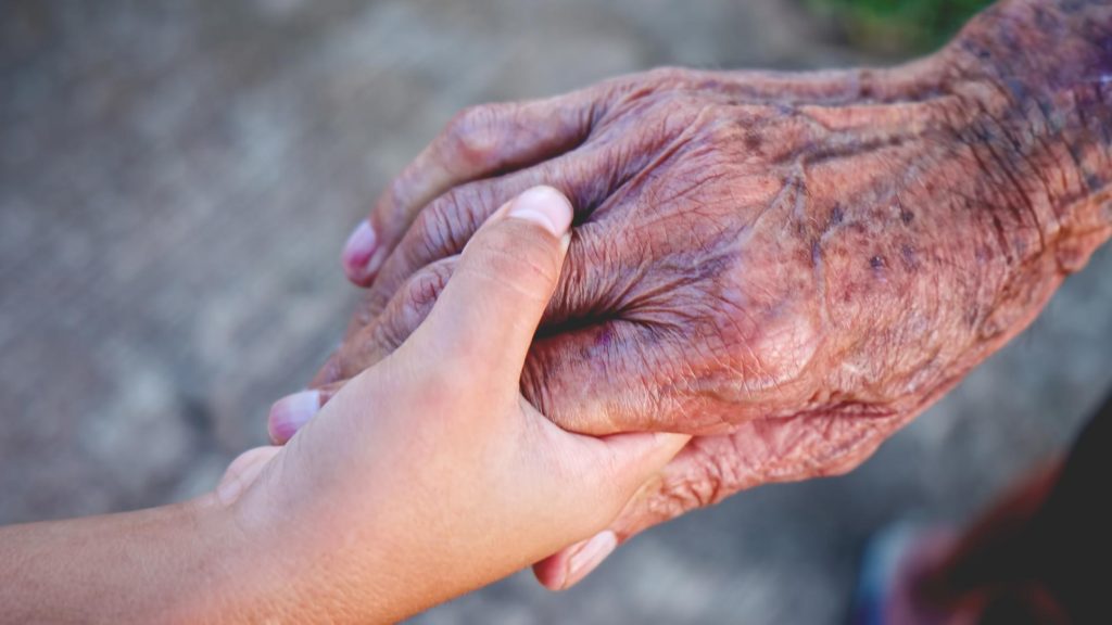 Photo of a younger female hand holding an older male hand with care
